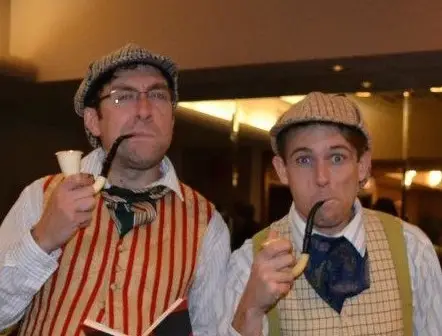 Two people wearing flat caps and vintage-style clothing, reminiscent of a classic murder mystery, hold pipes while looking at the camera. One sports a red-striped vest, the other green-striped, with scarves tied around their necks adding to their enigmatic charm.