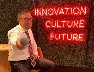 A man in business attire sits on a stool, holding a microphone toward the camera. Showcasing the expertise of Professional Emcees, a neon sign behind him reads "INNOVATION CULTURE FUTURE" against a vibrant green wall.