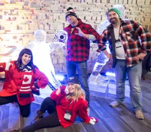 Four adults wearing red and plaid pose cheerfully in front of hockey player ice sculptures. Two stand and two kneel, all smiling, with Canadian-themed clothing and winter hats.