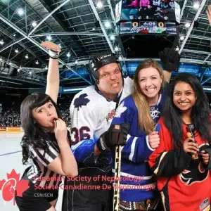 Four excited people pose at a hockey rink: a referee, a player in white gear, and two women in blue and red jerseys, all smiling and making playful gestures. The crowd and scoreboard are visible in the background.