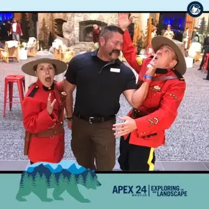 Three people pose playfully indoors: a man in black playfully holds back one person dressed as a Royal Canadian Mountie while another Mountie costume-wearer acts surprised. Event branding appears at the bottom.
