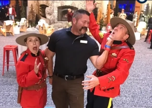 Three people pose playfully indoors: a man in black playfully holds back one person dressed as a Royal Canadian Mountie while another Mountie costume-wearer acts surprised. Event branding appears at the bottom.