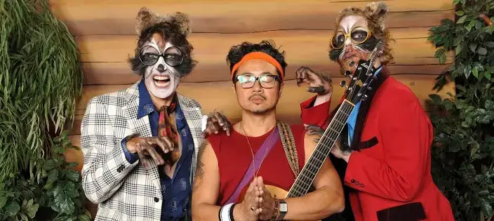 Three people pose together; two wear raccoon makeup, costumes, and glasses, while the person in the center wears a red headband, glasses, and holds their hands in a prayer gesture. One raccoon-costumed person holds a guitar.