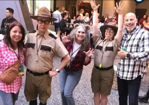Five smiling adults pose together indoors at an event. Two are dressed as park rangers in costume. The others wear casual clothes. "APEX 24 Whistler Exploring the Landscape" text with a bear and trees decorates the bottom.