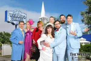 A group of people dressed in colorful, quirky outfits pose in front of a wedding chapel backdrop. The bride and groom are in the center, surrounded by friends in blue suits and vibrant dresses, smiling and making playful faces.