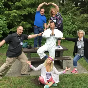 Five adults pose playfully around a picnic table in front of trees; one stands on the bench in a yoga pose, two make heart shapes above him, one points, and another sits cross-legged in front, all smiling and wearing casual clothes.