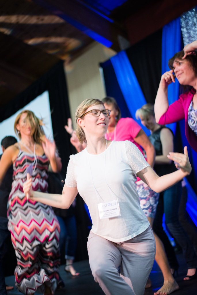 A group of people enthusiastically dancing indoors, with a woman in glasses and light clothing in the foreground, surrounded by others in colorful outfits—everyone radiates joy and energy as if celebrating after an inspiring keynote.