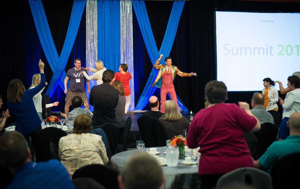 People at a conference watch and participate as two lively emcees, one in colorful overalls, lead an activity on stage. Blue and silver drapes decorate the backdrop, while a screen displays “Summit 201,” setting the scene for this engaging keynote session.