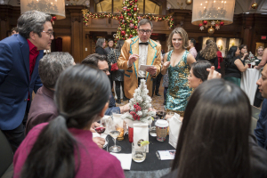 A group of people gather around a table at a festive holiday party, with a man in a gold jacket and a woman in a sequined dress chatting with guests. A decorated Christmas tree sparkles nearby as the group gets ready to bust a move on the dance floor.