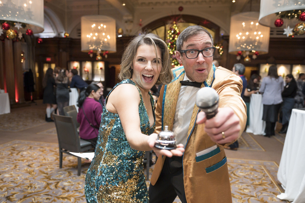 A woman in a sequined dress and a man in a gold jacket enthusiastically pose together, ready to bust a move with a desk bell and microphone, at a festive indoor event featuring holiday decorations and sparkling chandeliers.