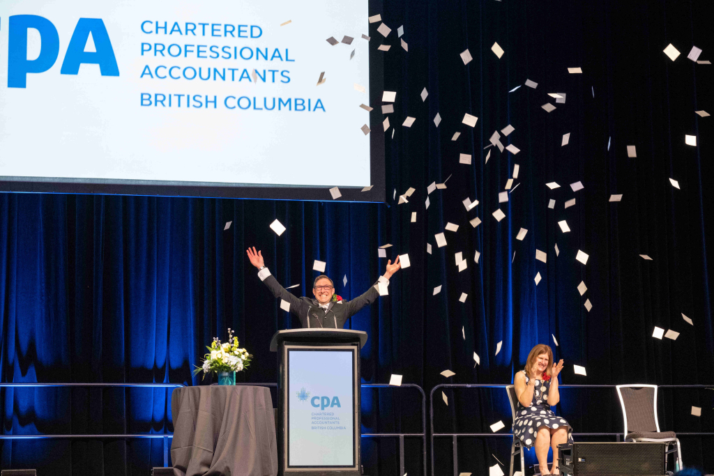 A person stands at a podium with arms raised and papers flying in the air, while another person sits and claps on stage. A large CPA British Columbia sign is displayed above them.