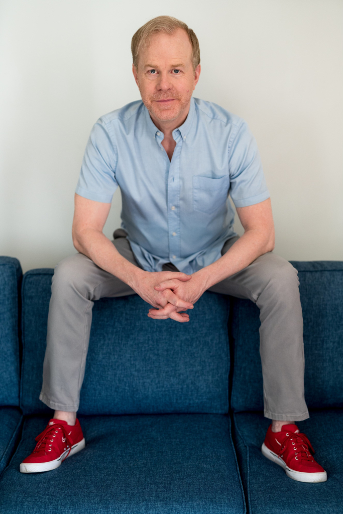 A man with short blond hair, wearing a light blue shirt, gray pants, and red sneakers, sits on the backrest of a blue sofa, leaning forward with hands clasped and looking at the camera—capturing the confident poise of professional emcees.
