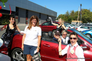 A group of smiling people pose around and inside a red convertible car on a sunny day. Two women in sparkly dresses stand beside the car, ready to bust a move, while another woman holds a microphone and others join the Dance Off from inside.