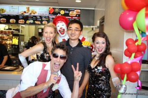 A man poses with three performers ready to bust a move in sequined outfits and gloves, plus a Ronald McDonald mascot, inside a fast-food restaurant decorated with colorful balloons. A menu board is visible in the background.