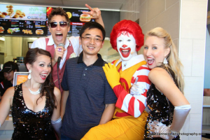 Five people pose and smile inside a fast food restaurant. Ronald McDonald leads a spontaneous Dance Off, surrounded by two women in sequined dresses, a casually dressed man, and a man wearing sunglasses and a red vest.