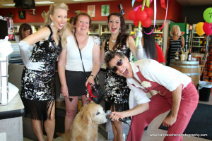 Four adults pose and smile in a liquor store; two women in black sequined dresses look ready to bust a move, while another woman in casual clothes and a man in red suspenders kneels to feed a treat to a golden retriever. Red balloons hang in the background.