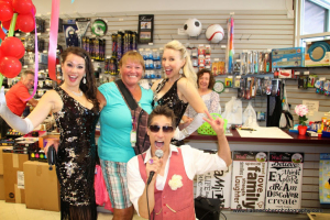 A woman stands in a store, smiling with three performers in sequined outfits; one man sings into a microphone while the others pose with balloons, ready to bust a move. The background features shelves of colorful items and vibrant wall decor.