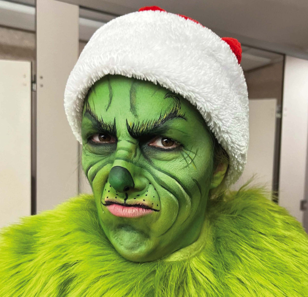 Person dressed as the Grinch, with green face paint, detailed black lines, a Santa hat, and a furry green costume, making a grumpy facial expression.