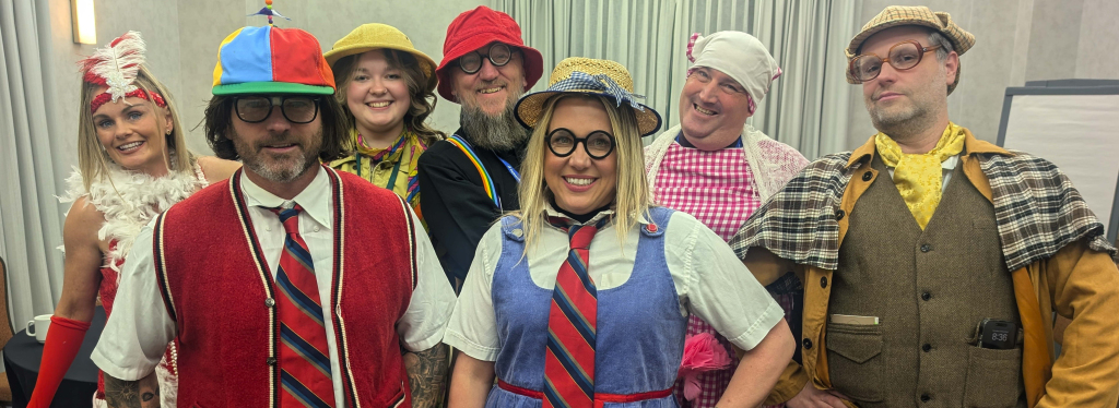Seven adults dressed in colorful, playful costumes and hats, smiling at the camera in a room with grey curtains. Their outfits feature stripes, checks, ties, and accessories for a cheerful group scene fit for entertainment news.