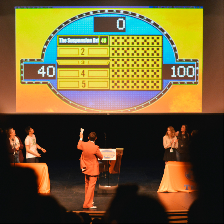 A person in an orange suit hosts a Family Feud-style game on stage with two teams standing at orange tables, while a large game board showing scores is projected behind them.
