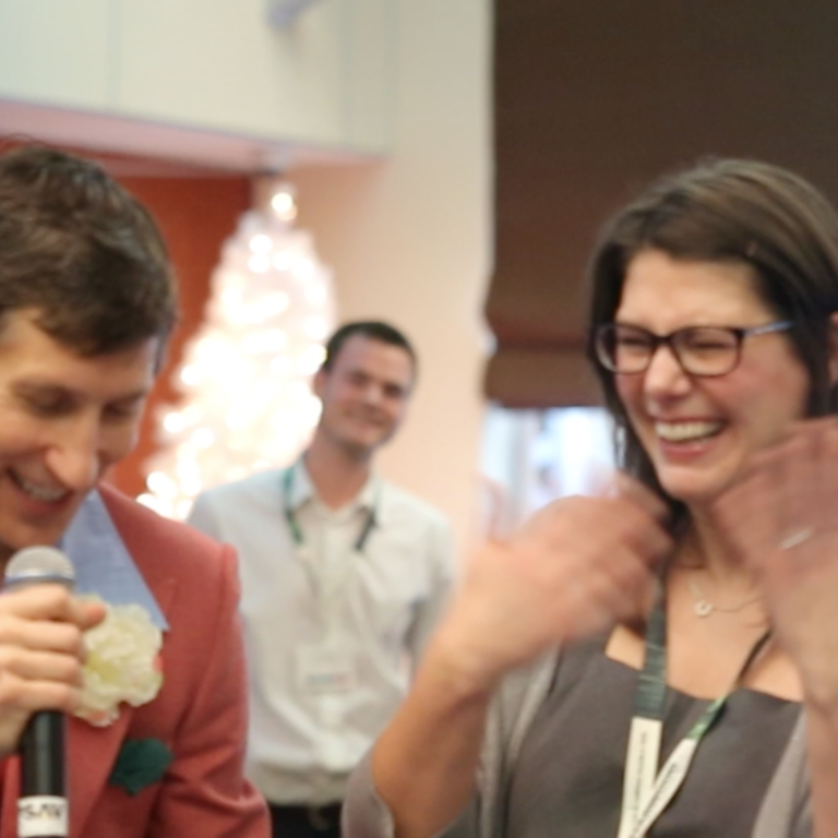 Two people stand together, smiling and laughing. One holds a microphone and wears a suit with a boutonniere. Behind them, a blurred person in a white shirt and a lit, decorative tree are visible in the background.