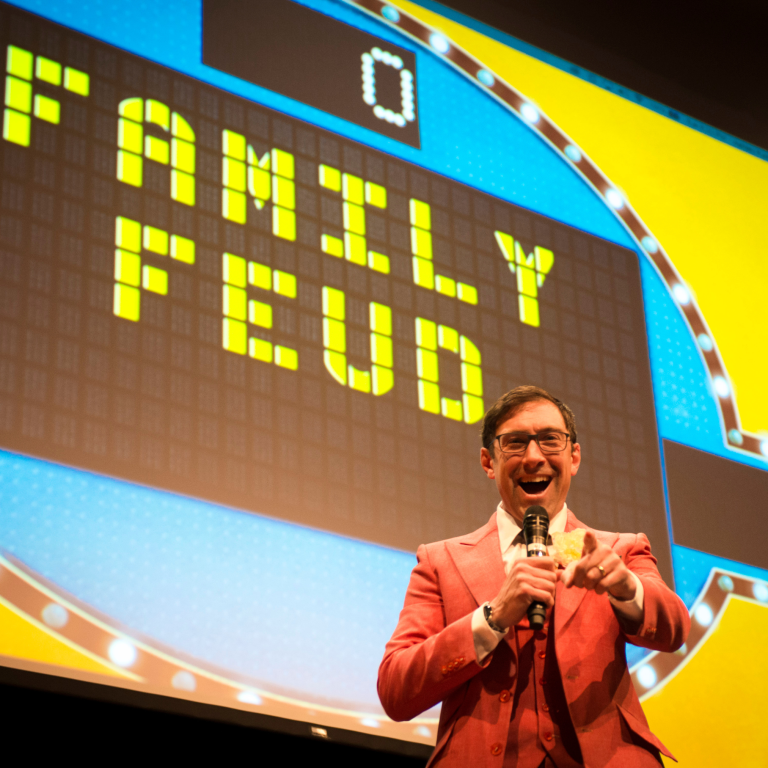 A game show host in a red suit holds a microphone and smiles while pointing at the audience, standing in front of a large Family Feud game board display with a score of zero.