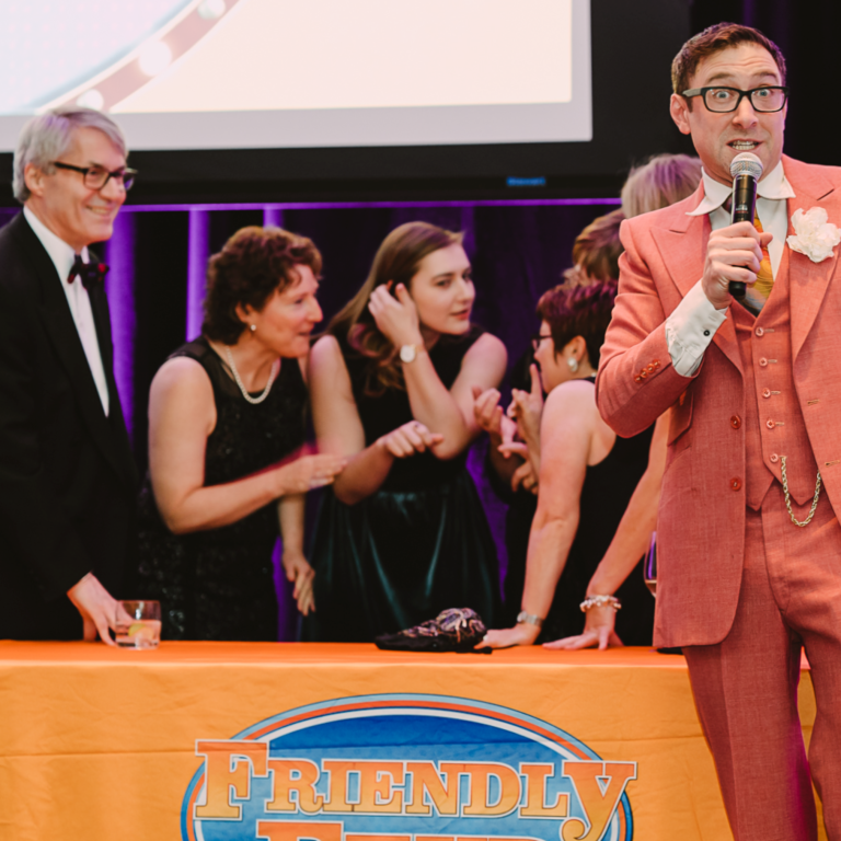 A man in a coral suit speaks into a microphone at a podium labeled "Friendly," while a group of smiling people stand behind the table, appearing engaged and excited at an indoor event.
