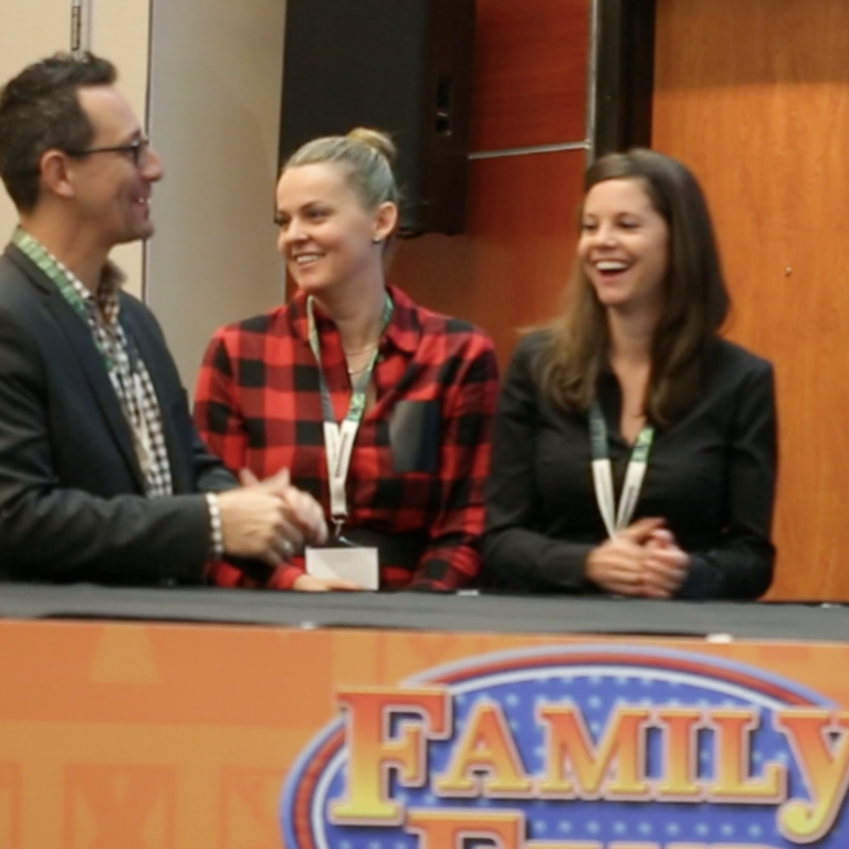 Three people sit at a table, smiling and laughing during what appears to be a panel or event. Two women sit side by side, while a man faces them and gestures with his hands. A partial "Family" sign is visible in front.