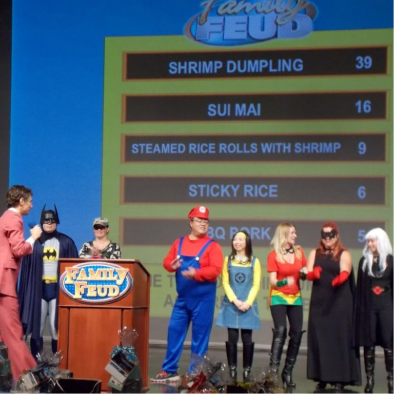 A group of people in various costumes stand on stage during a "Family Feud" game show, with a host in a pink suit. A scoreboard behind them displays Asian food items and point values.