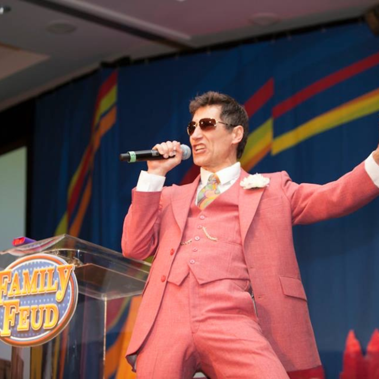 A man in a pink suit and sunglasses holds a microphone and gestures energetically on a stage with a "Family Feud" sign in front of him. The background features colorful lines on a blue curtain.