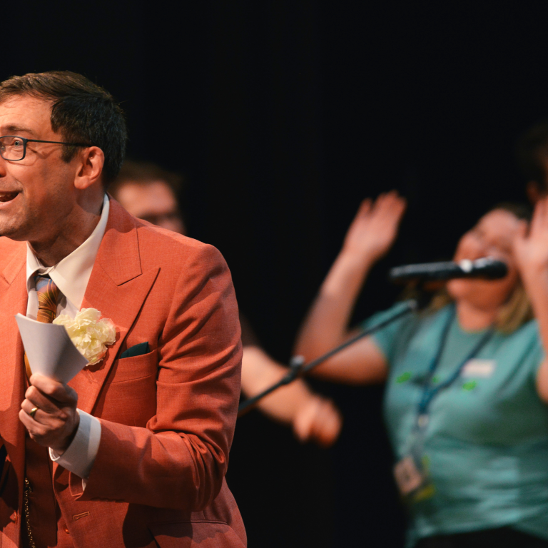 A man in a salmon-colored suit and patterned tie speaks enthusiastically on stage, holding a pen. Behind him, a woman sings into a microphone with raised hands, and other people are visible in the background.