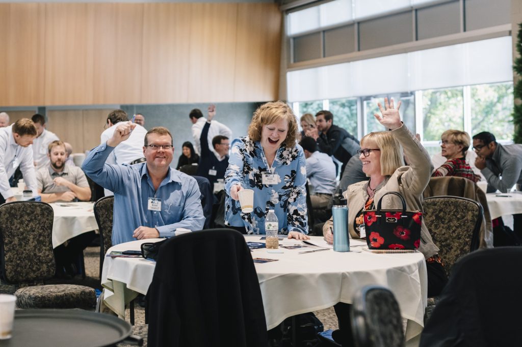 Three people sit at a round table during a conference, smiling and raising their hands to boost engagement. Other attendees are visible in the background, all seated in a bright room with large windows.
