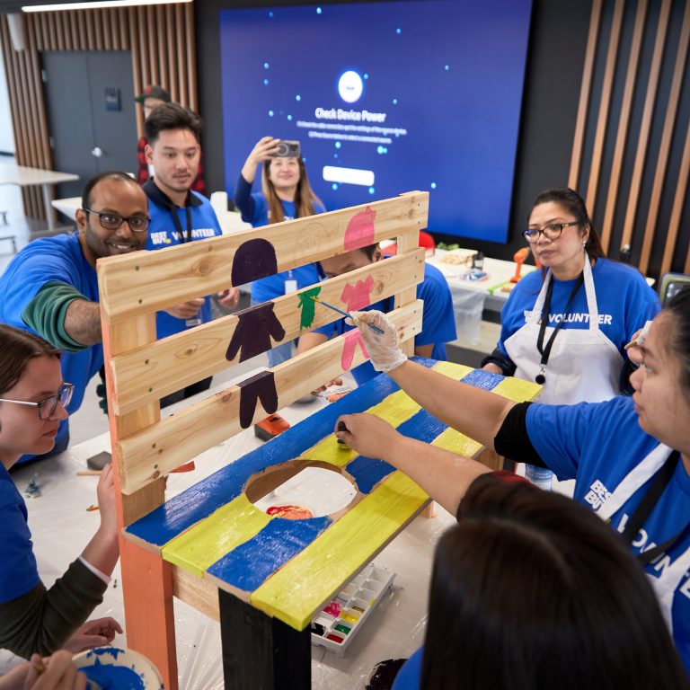 A group of volunteers in blue shirts paint a wooden bench with colorful designs, including silhouettes of children, at an indoor event. One person takes a photo while others focus on decorating the bench together.