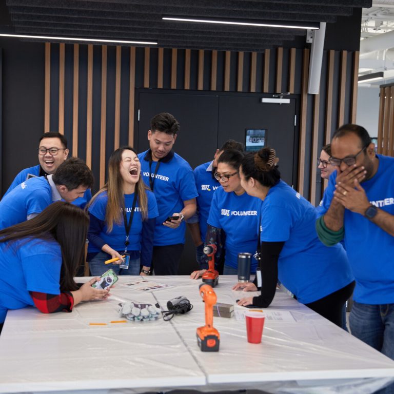 A group of people in blue volunteer shirts stand and laugh around a table with tools and papers, appearing to enjoy a collaborative activity in a modern indoor space.