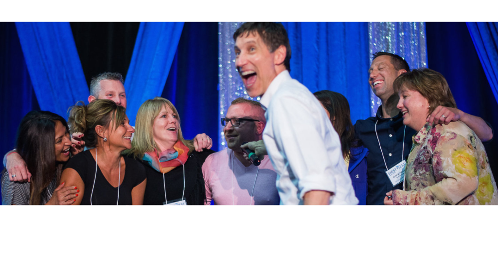 Roger on stage with laughing group First West cropped 1200 x 630 px