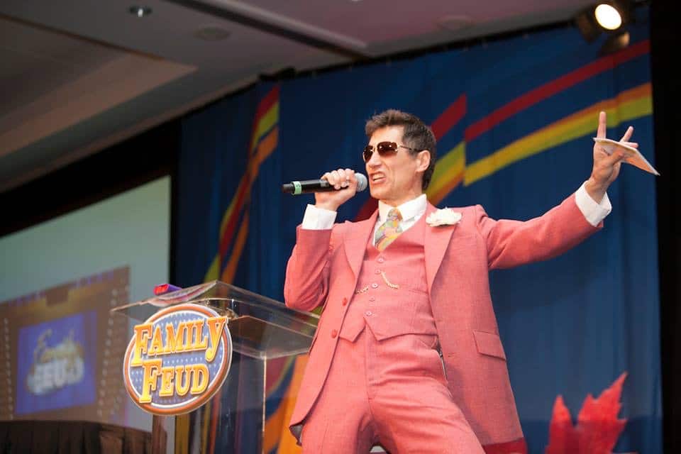 Roger, in character as a flashy game-show host, fires up the crowd behind the Family Feud podium.