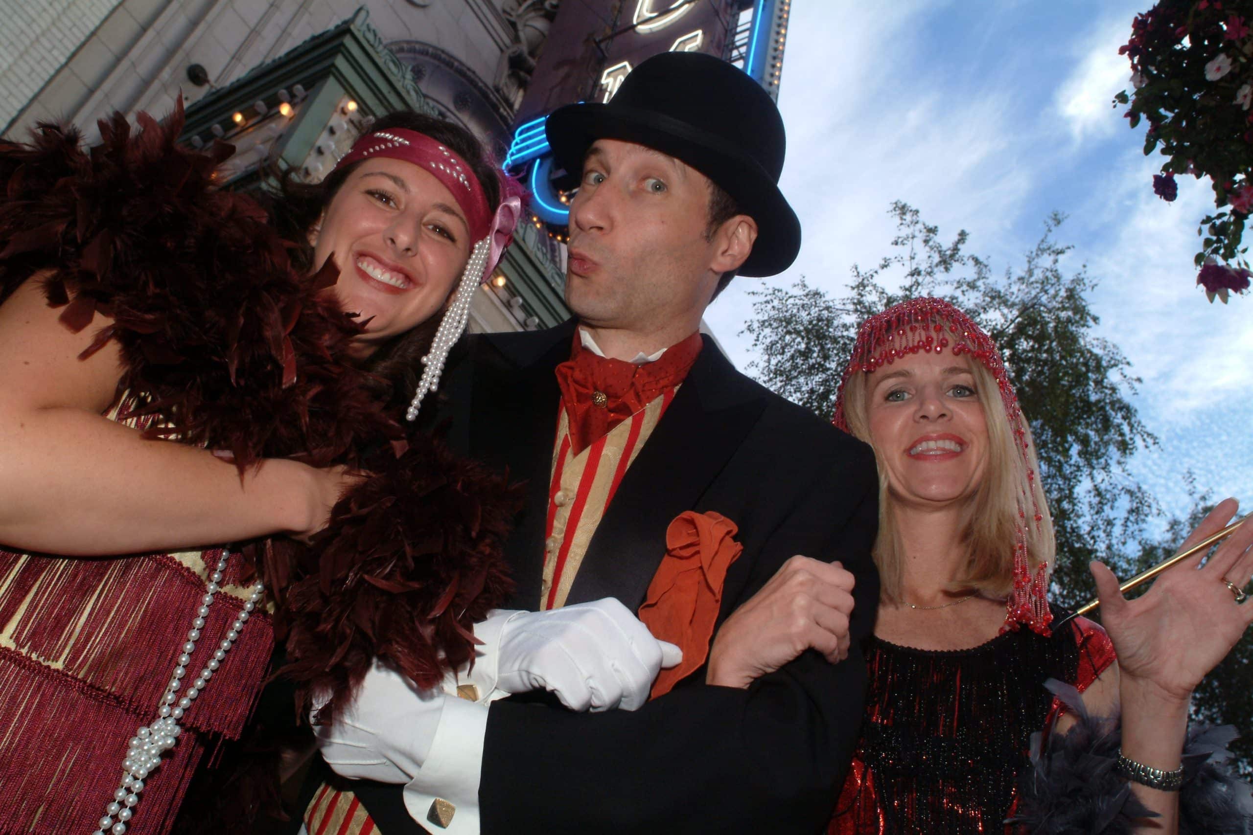 Roger in 1920s Gatsby-style attire poses with two flapper-costumed guests outside a theatre marquee.