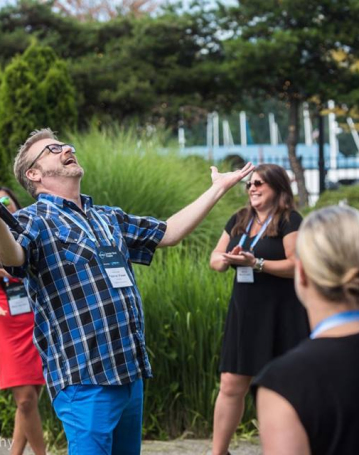 A man in a blue plaid shirt stands outdoors with arms outstretched, smiling joyfully, as a group of people enjoy the entertainment around him. Lush greenery and trees create a vibrant backdrop.