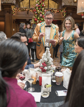 A festive group gathers around a table at a holiday party, enjoying lively entertainment. A man in a gold jacket and a woman in a sparkly teal dress stand, engaging others. Decorated Christmas trees and ornaments enhance the elegant background.