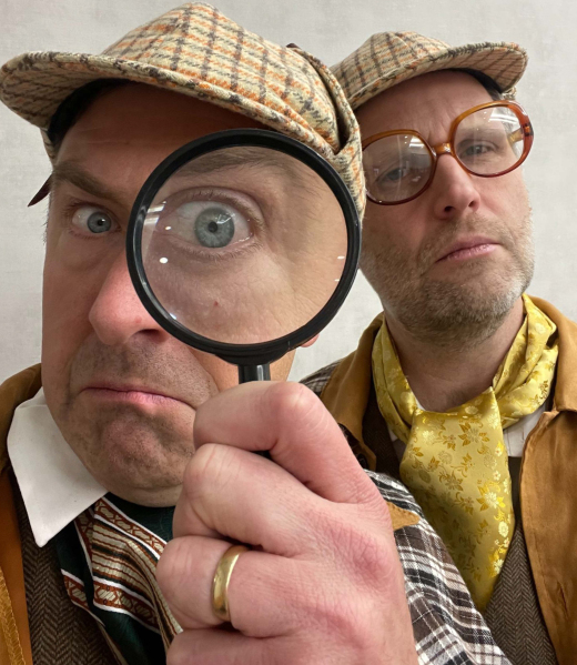 Two men dressed as detectives in vintage clothing—one with a magnifying glass and checkered hat, the other in glasses and a yellow patterned scarf—stare seriously at the camera, ready to crack the latest entertainment news mystery.